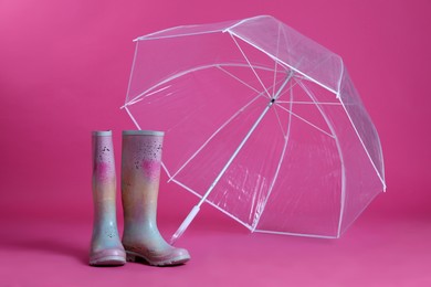 Photo of Open transparent umbrella and rubber boots on pink background