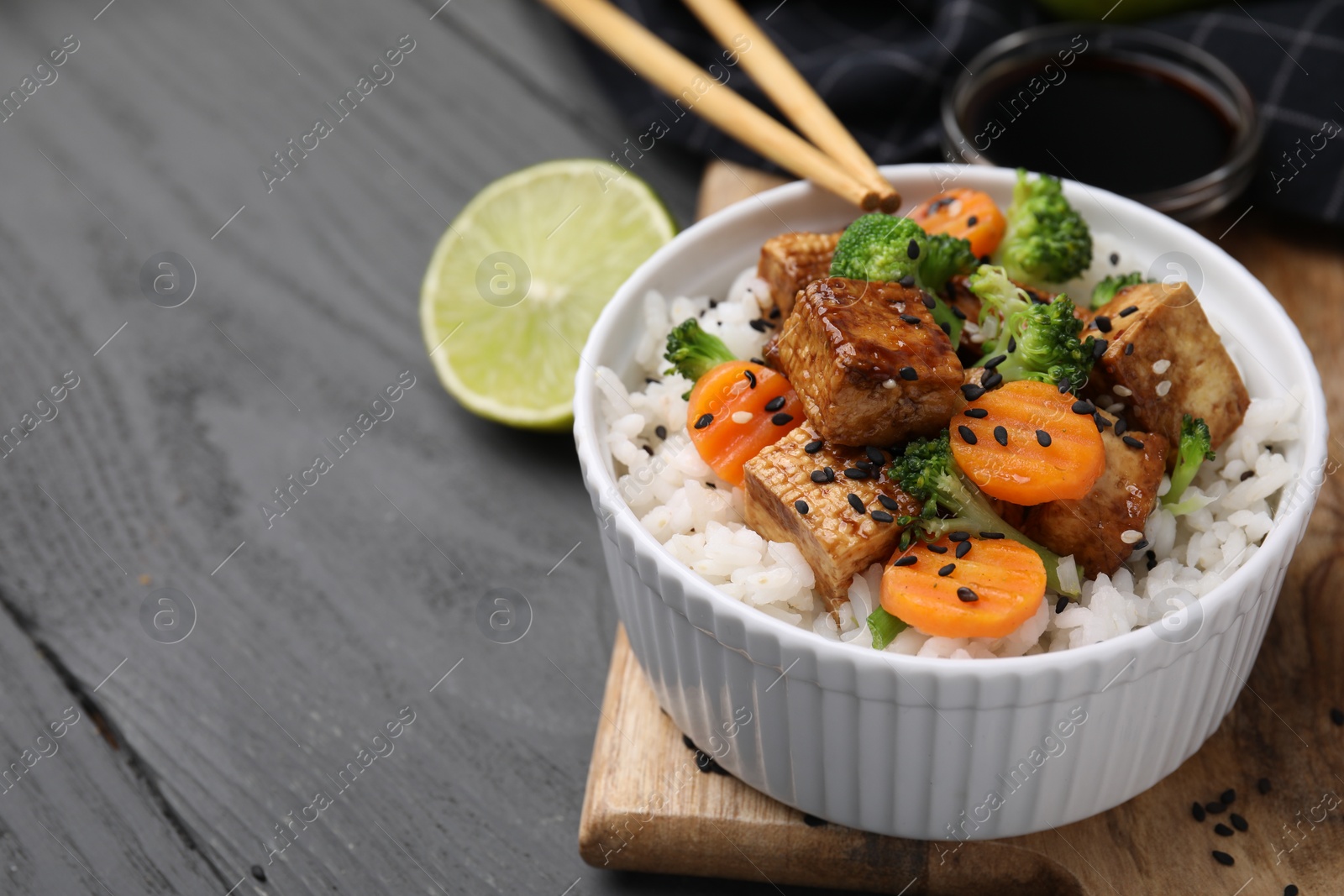 Photo of Bowl of rice with fried tofu, broccoli and carrots on grey wooden table, closeup. Space for text