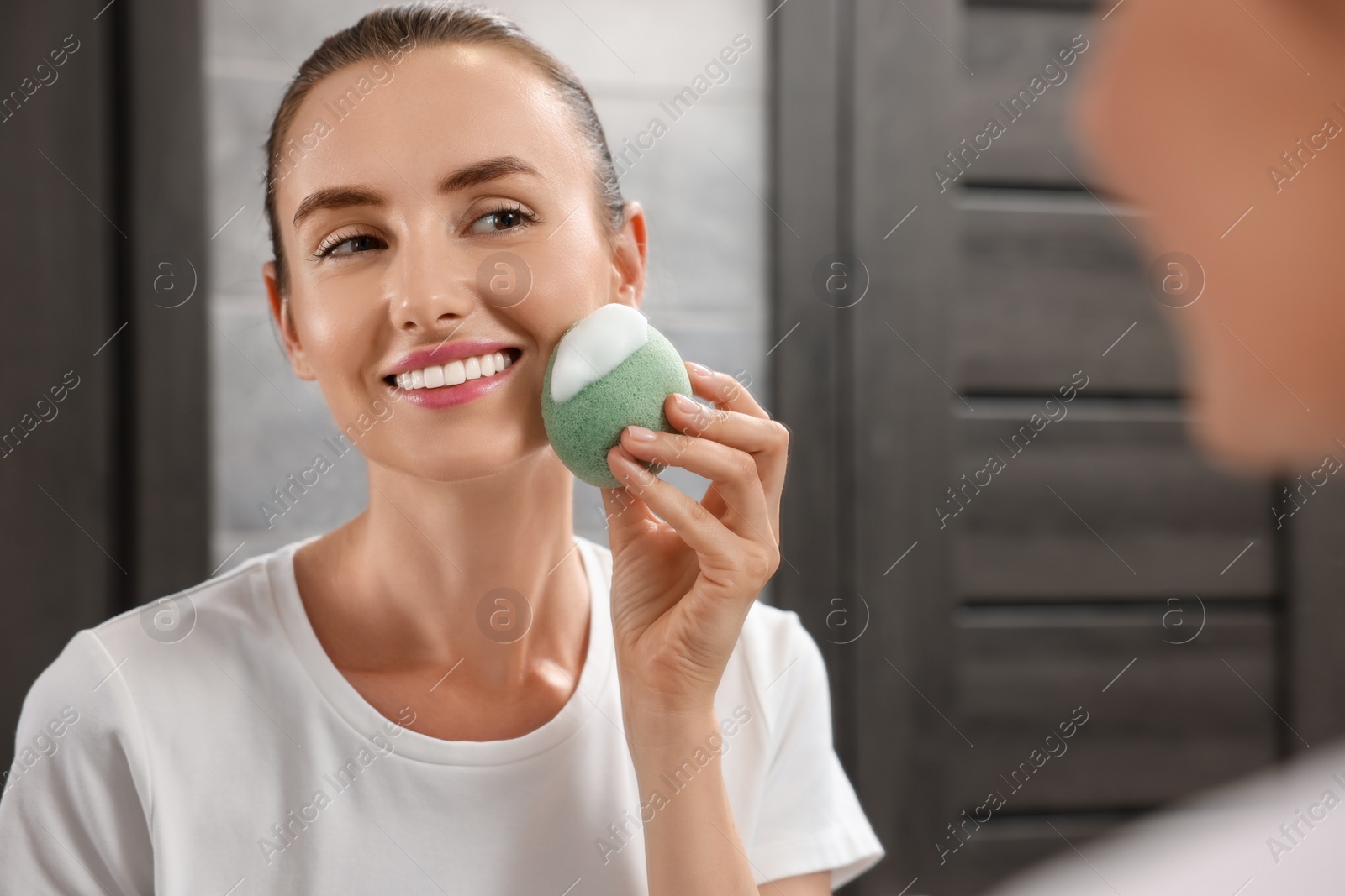 Photo of Happy young woman washing her face with sponge near mirror in bathroom