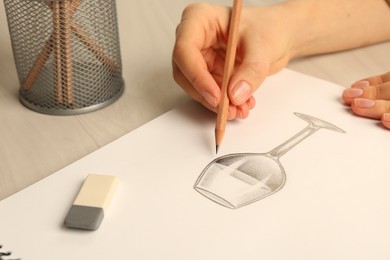 Photo of Woman drawing glass of wine with graphite pencil at light wooden table, closeup