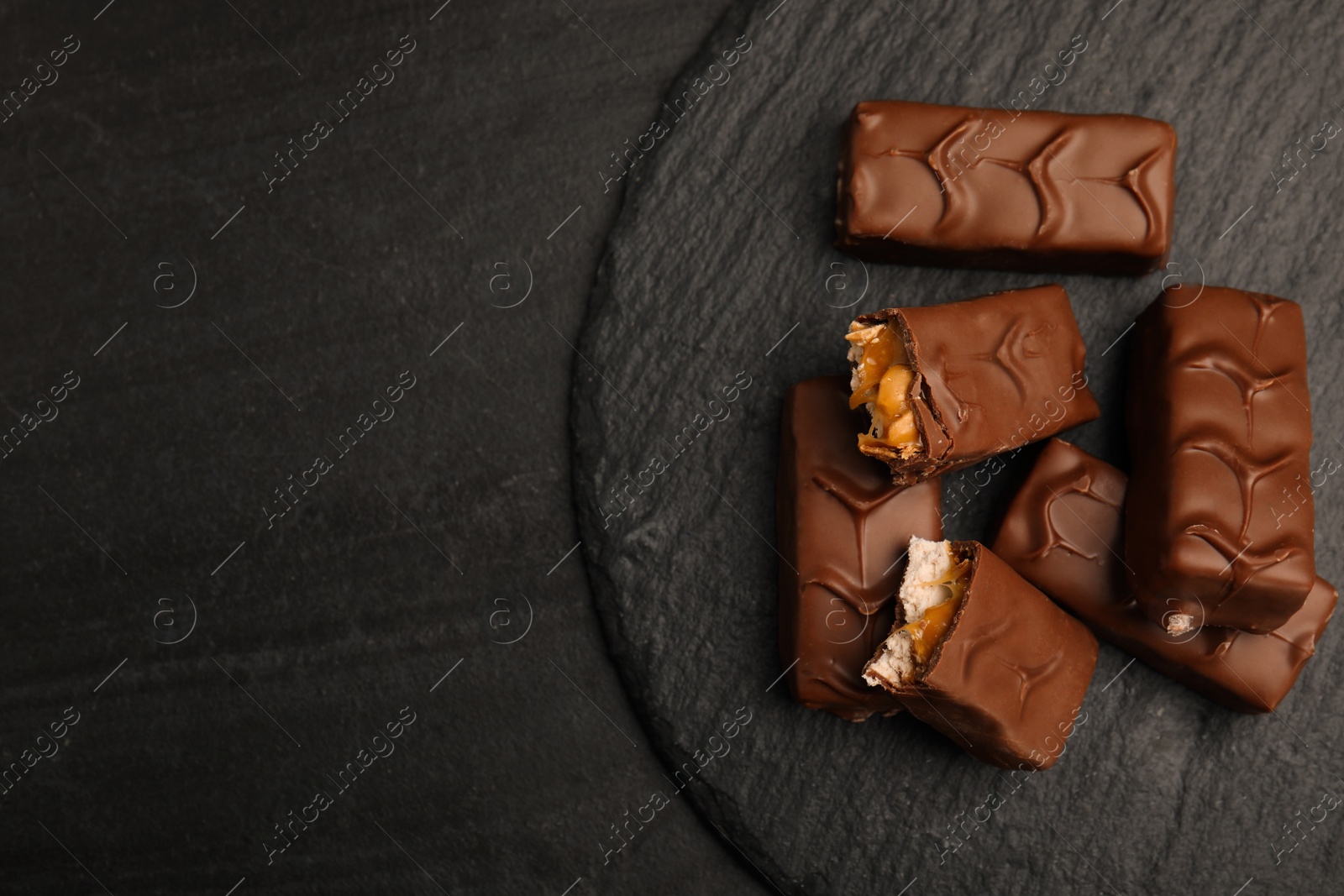 Photo of Slate plate of chocolate bars with caramel, nuts and nougat on black table, top view. Space for text