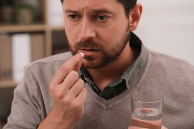 Depressed man with glass of water taking antidepressant pill on blurred background, closeup