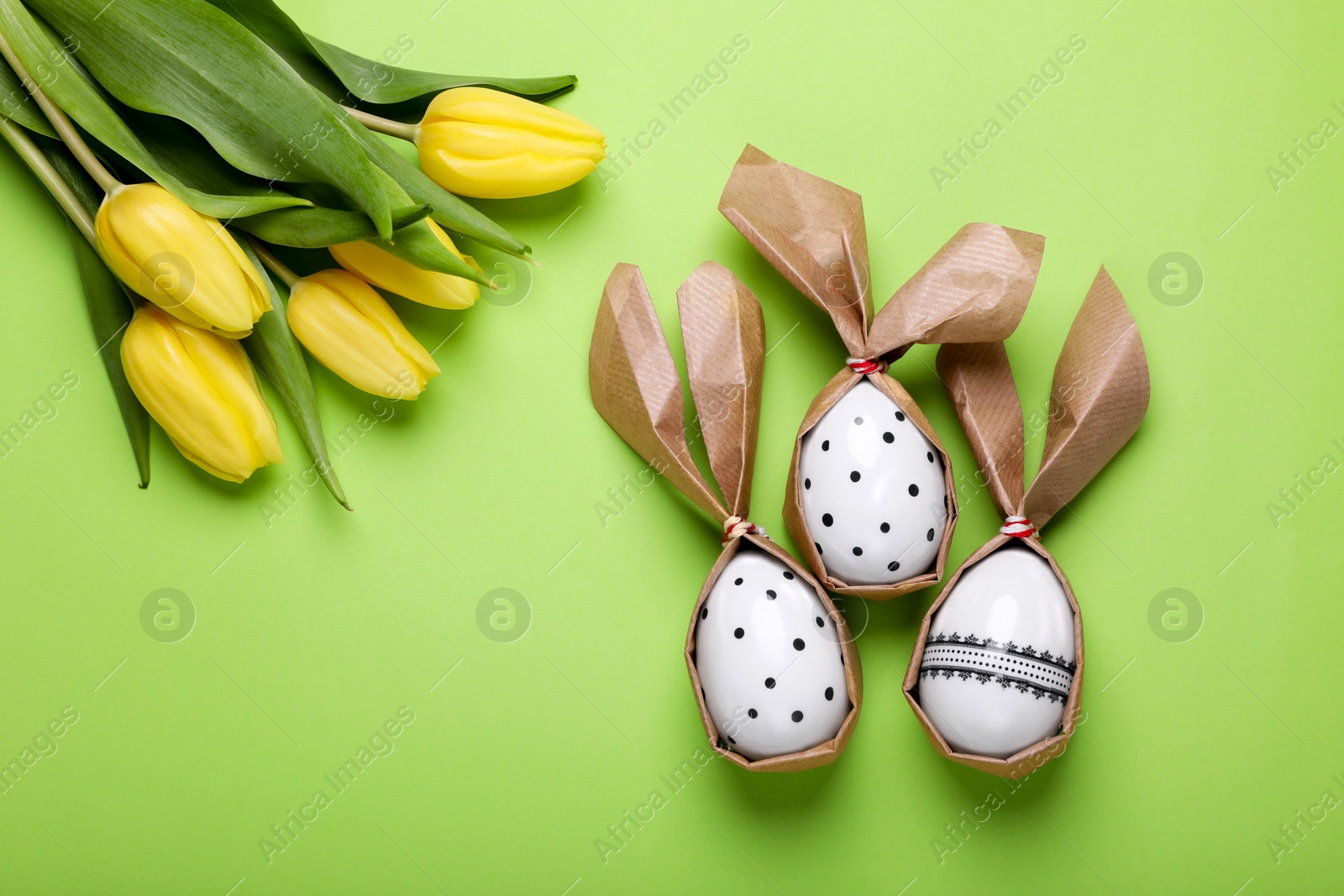 Photo of Easter bunnies made of craft paper and eggs near beautiful tulips on light green background, flat lay