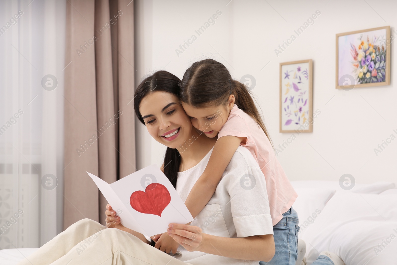 Photo of Happy woman with her daughter and handmade greeting card on bed at home. Mother's day celebration