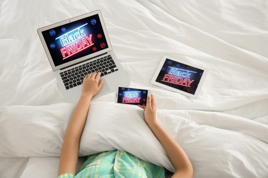 Photo of Woman using tablet, mobile phone and laptop with Black Friday announcement while lying in bed, above view
