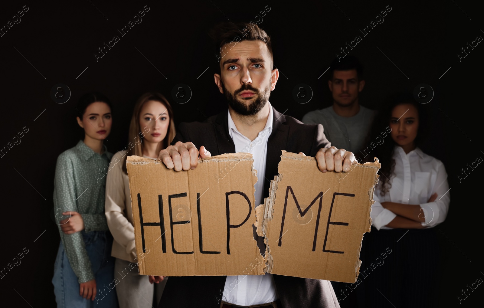 Photo of Unhappy man with HELP ME sign and group of people behind his back on dark background
