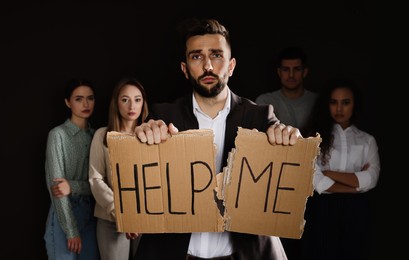 Photo of Unhappy man with HELP ME sign and group of people behind his back on dark background