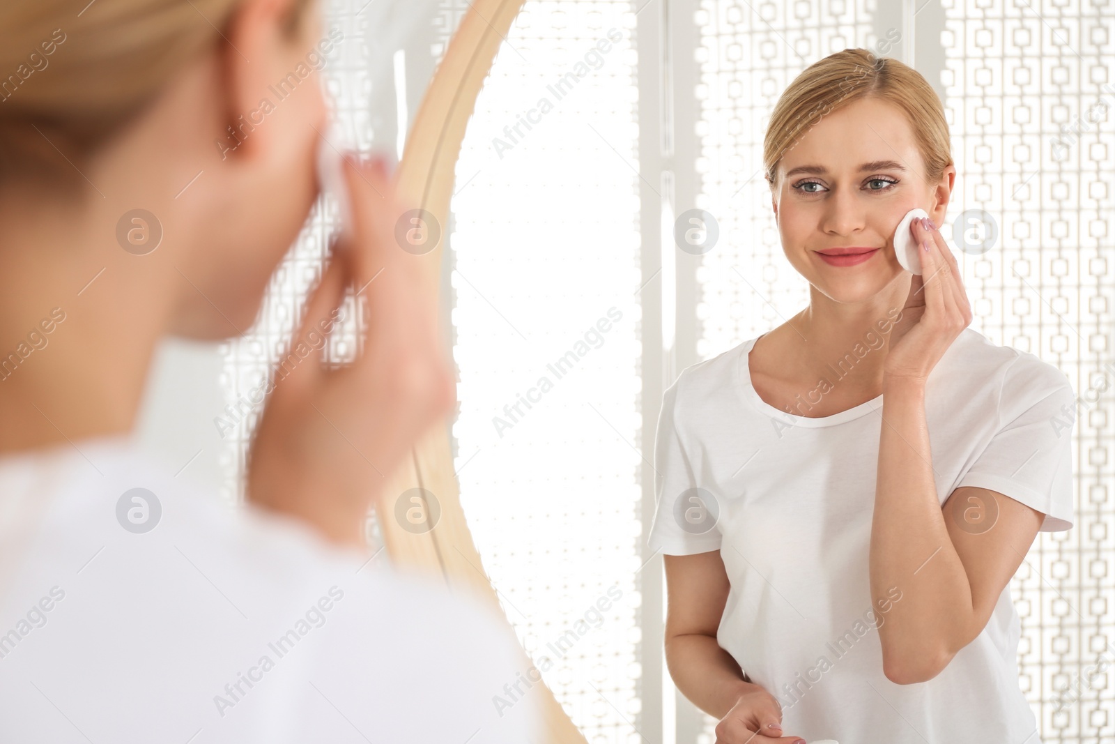 Photo of Happy young woman cleaning face with cotton pad near mirror in bathroom