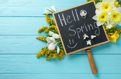 Photo of Blackboard with words HELLO SPRING and flowers on light blue wooden table, flat lay