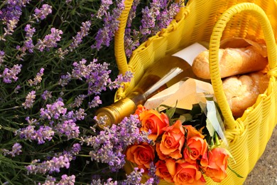 Photo of Yellow wicker bag with beautiful roses, bottle of wine and baguettes near lavender flowers outdoors, above view