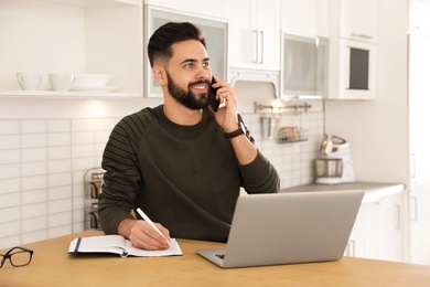 Photo of Handsome young man talking on phone while working at table in kitchen