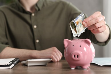 Photo of Financial savings. Man putting dollar banknote into piggy bank at wooden table, closeup