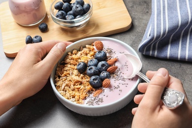 Woman eating tasty yogurt with oatmeal and blueberries at grey table, closeup