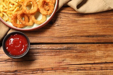 Photo of Tasty fried onion rings and french fries with ketchup on wooden table, flat lay. Space for text