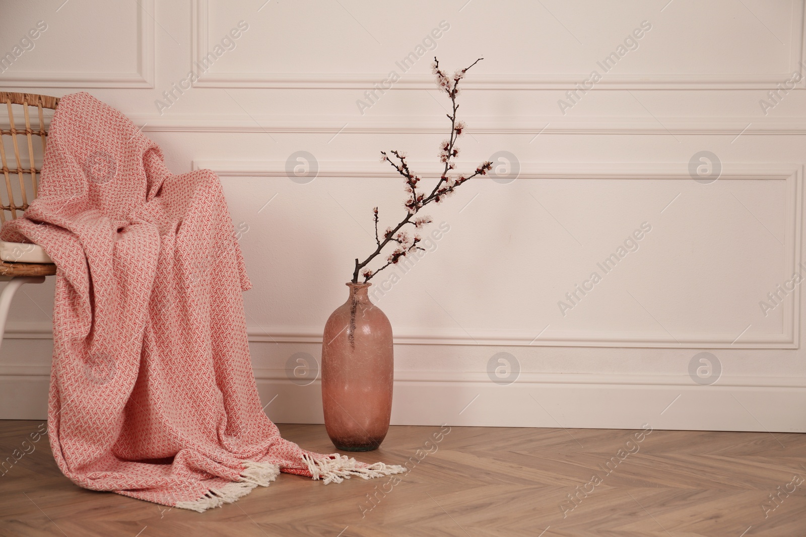 Photo of Flowering tree twig in glass vase on floor near chair with blanket at white wall. Space for text