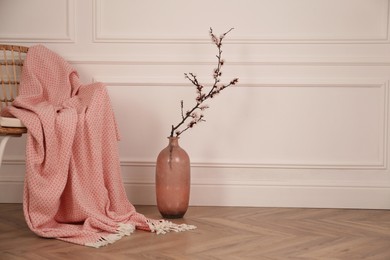 Photo of Flowering tree twig in glass vase on floor near chair with blanket at white wall. Space for text