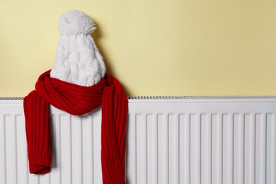 Knitted hat and scarf on heating radiator near beige wall, space for text