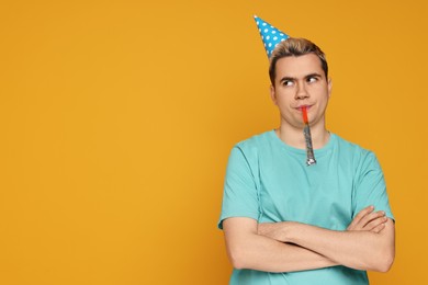 Sad young man with party hat and blower on orange background, space for text