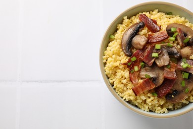 Photo of Tasty millet porridge with mushrooms, bacon and green onion in bowl on white tiled table, top view. Space for text