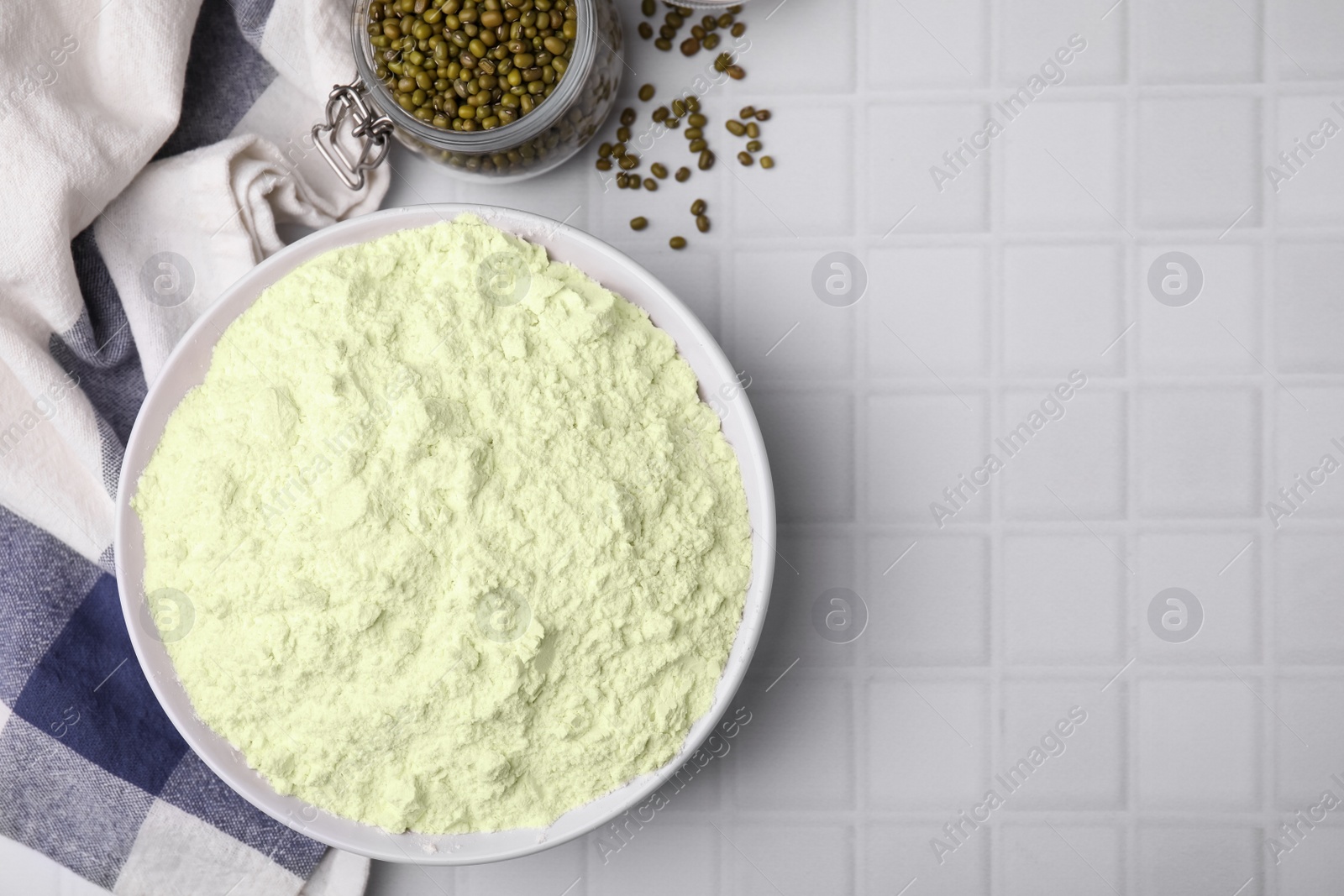 Photo of Mung bean flour in bowl and seeds on white tiled table, flat lay. Space for text