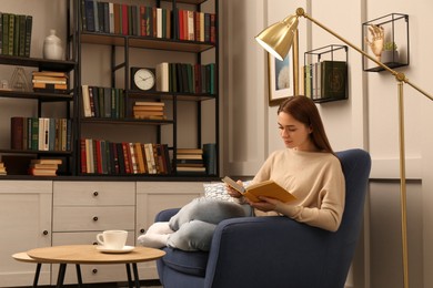 Young woman reading book in armchair indoors. Home library