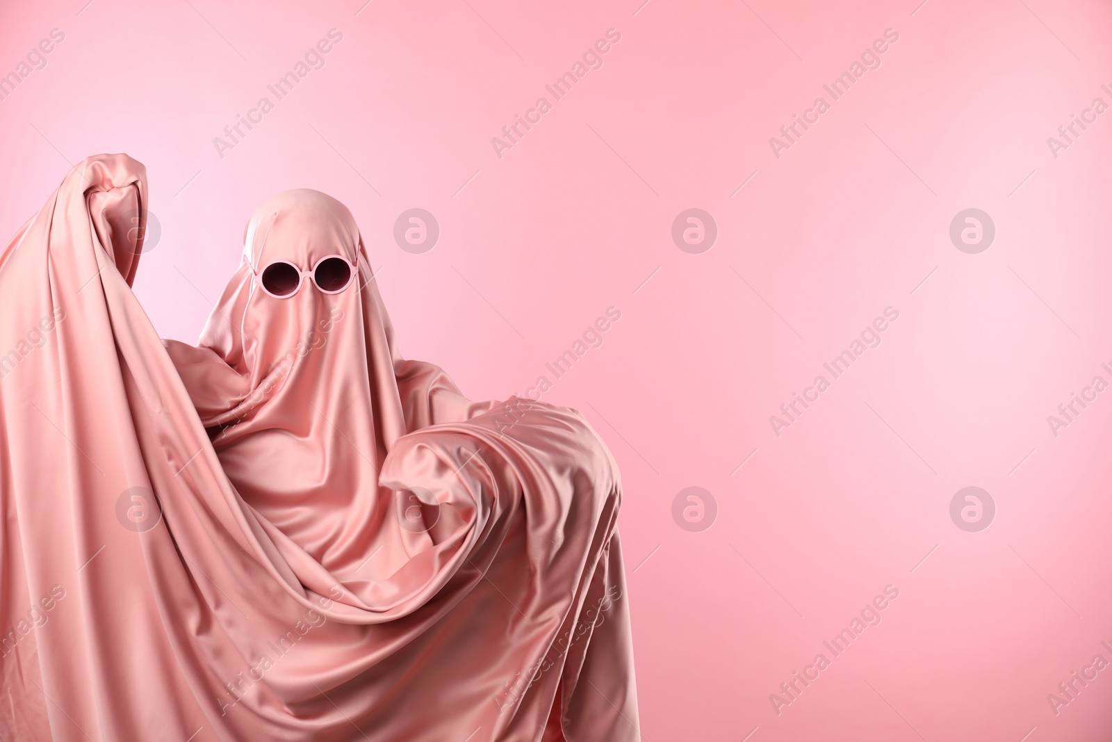 Photo of Glamorous ghost. Woman in sheet with sunglasses on pink background, space for text