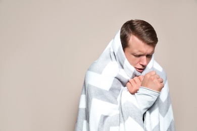 Image of Man wrapped in blanket coughing on beige background, space for text. Cold symptoms