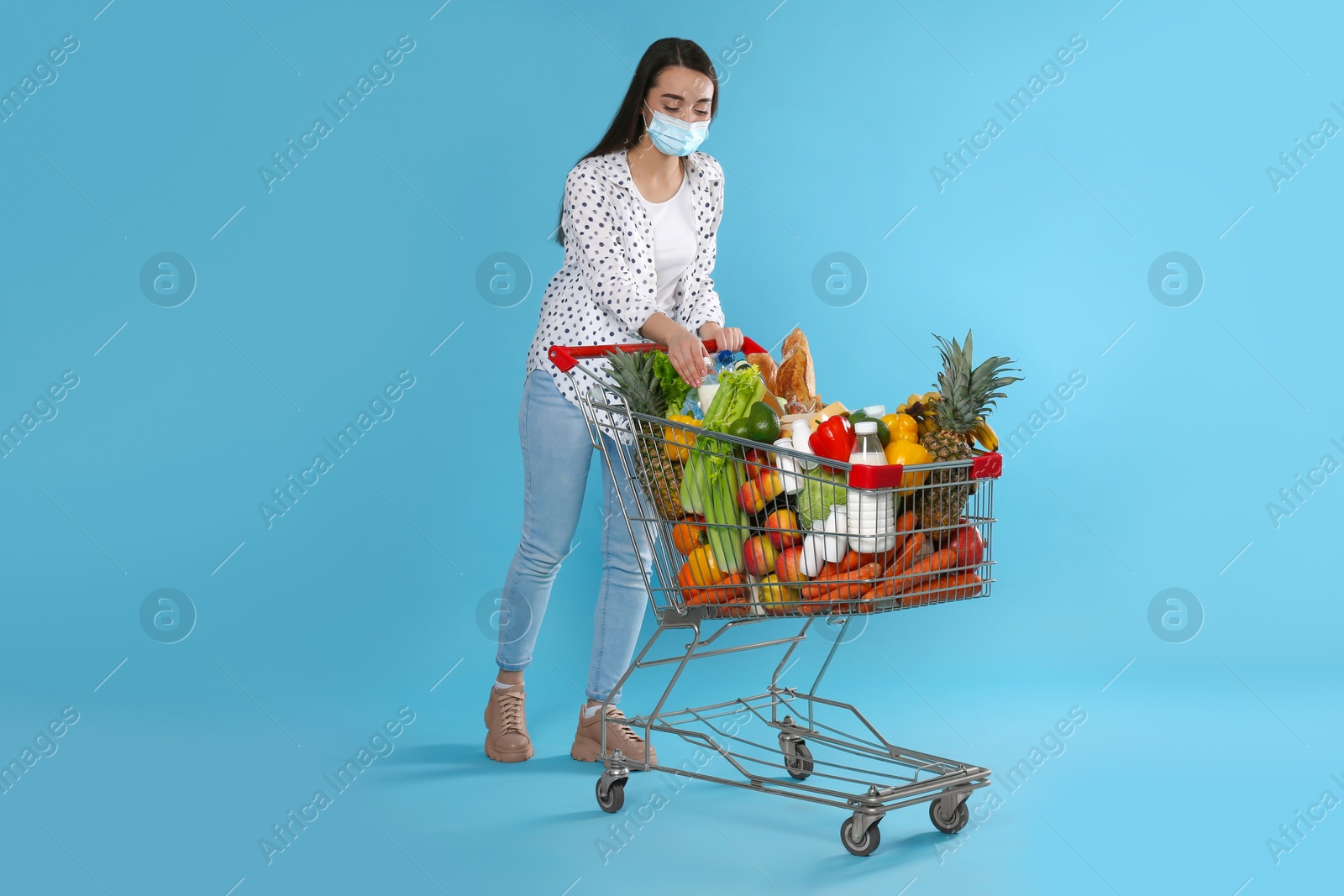 Photo of Young woman in medical mask with shopping cart full of groceries on light blue background