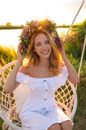 Young woman wearing wreath made of beautiful flowers on swing chair outdoors at sunset