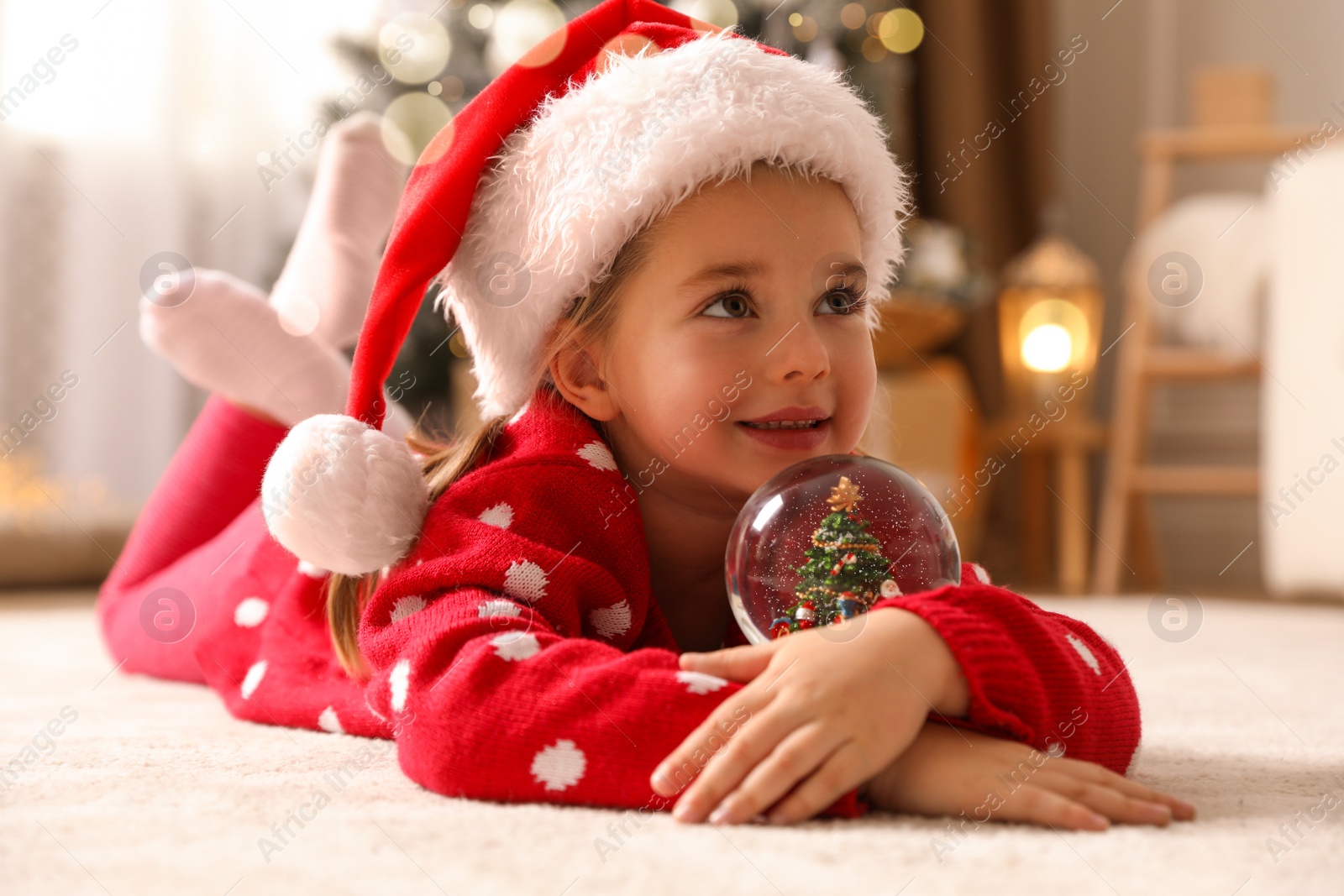 Photo of Little girl in Santa hat playing with snow globe on floor