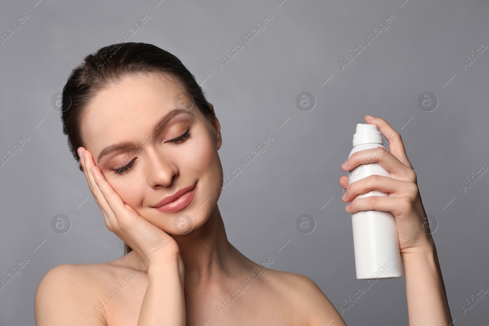 Photo of Young woman applying thermal water on face against grey background. Cosmetic product