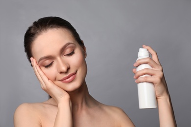 Young woman applying thermal water on face against grey background. Cosmetic product