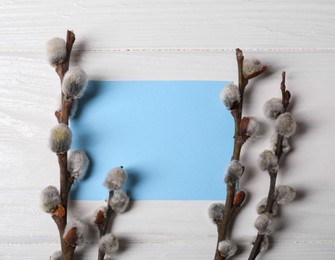 Beautiful blooming willow branches and blank card on white wooden table, flat lay. Space for text