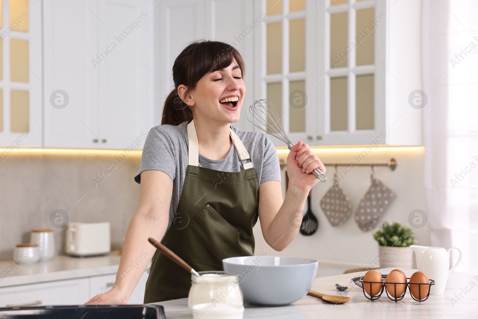 Photo of Happy young housewife with whisk having fun while cooking at white marble table in kitchen