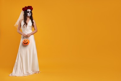 Photo of Young woman in scary bride costume with sugar skull makeup, flower crown and pumpkin bucket on orange background, space for text. Halloween celebration