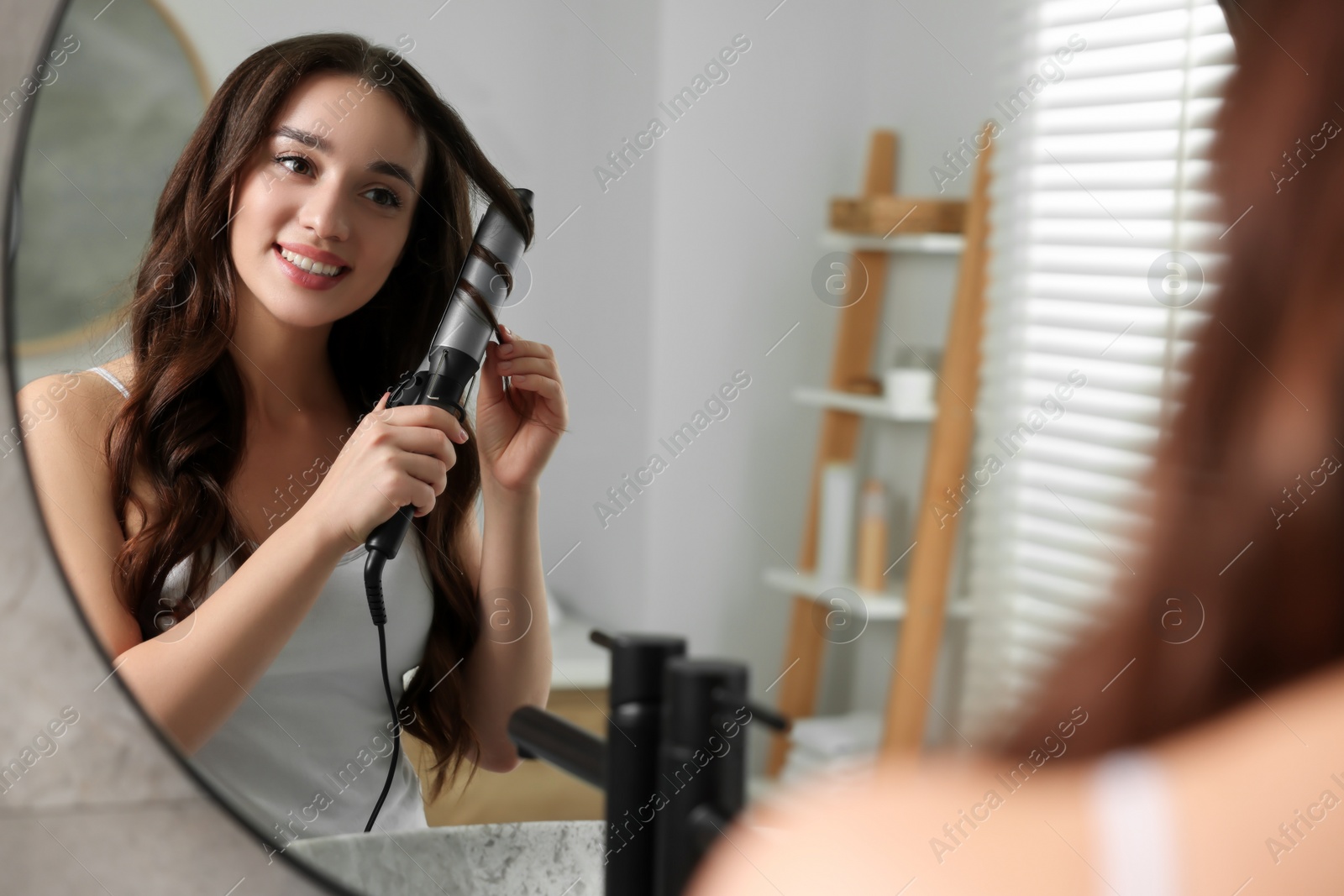 Photo of Smiling woman using curling hair iron near mirror in bathroom