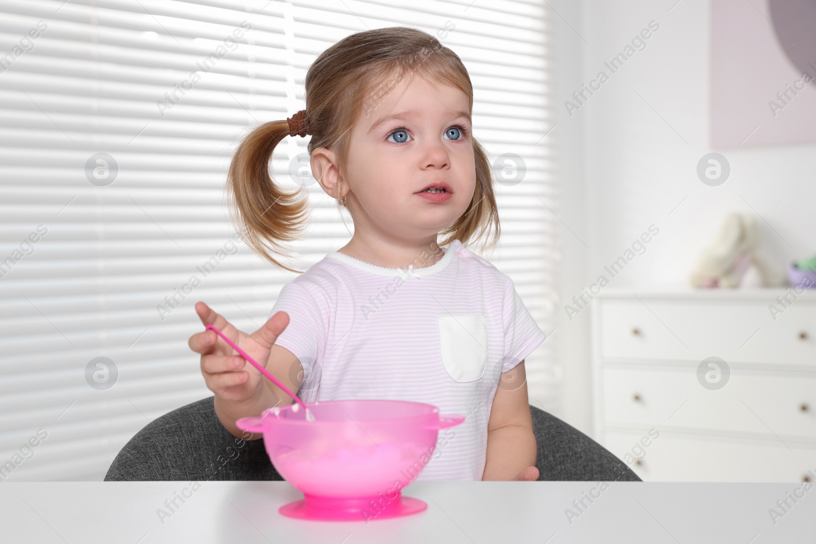 Photo of Cute little child eating tasty yogurt from plastic bowl with spoon at white table indoors