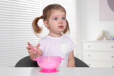 Cute little child eating tasty yogurt from plastic bowl with spoon at white table indoors