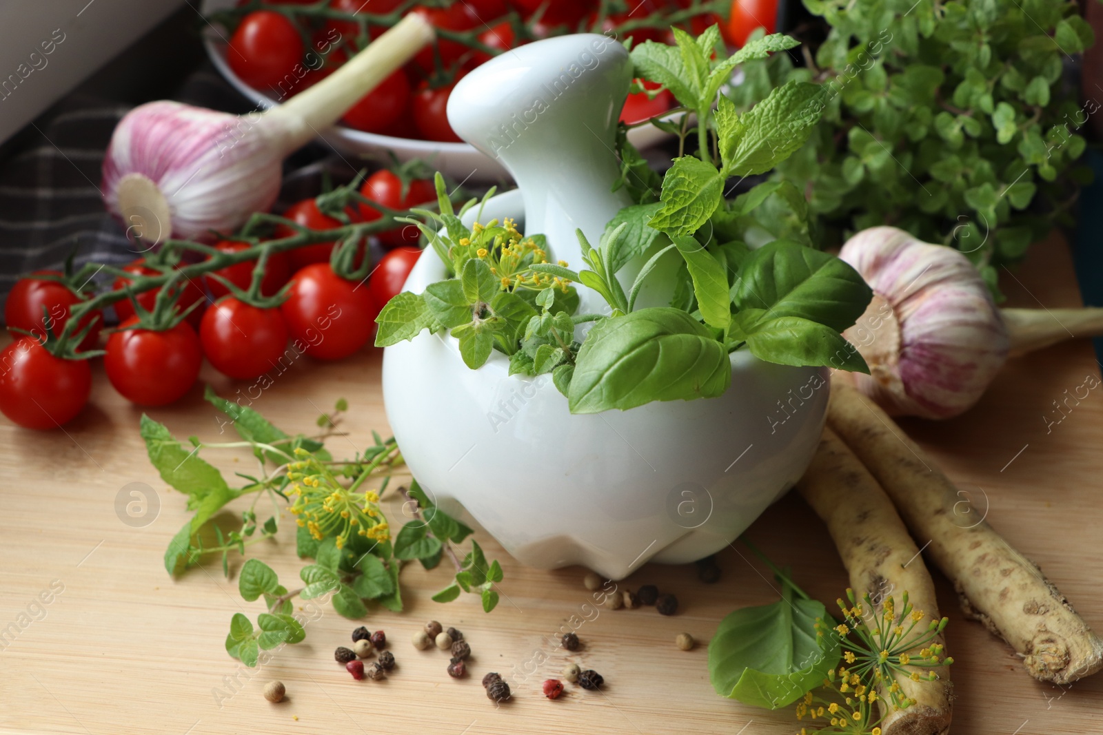 Photo of Mortar with fresh herbs near garlic, horseradish roots, black peppercorns and cherry tomatoes on wooden table, closeup