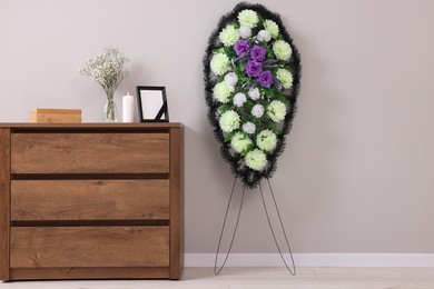 Photo of Wreath of plastic flowers and frame with black ribbon, burning candle on commode in room. Funeral attributes