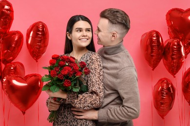 Happy couple celebrating Valentine's day. Beloved woman holding bouquet of roses near heart shaped air balloons on red background