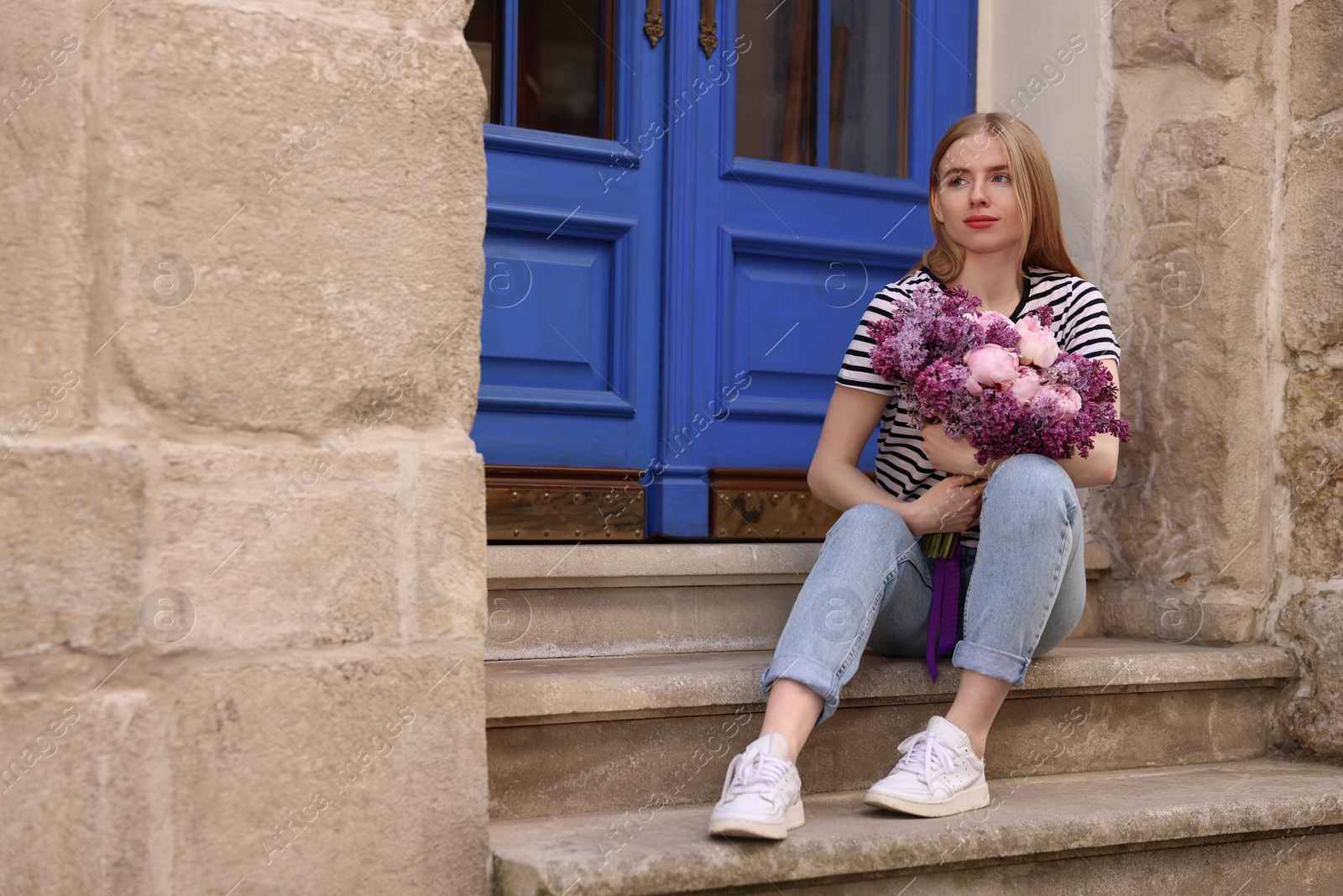 Photo of Beautiful woman with bouquet of spring flowers on stairs near building, space for text