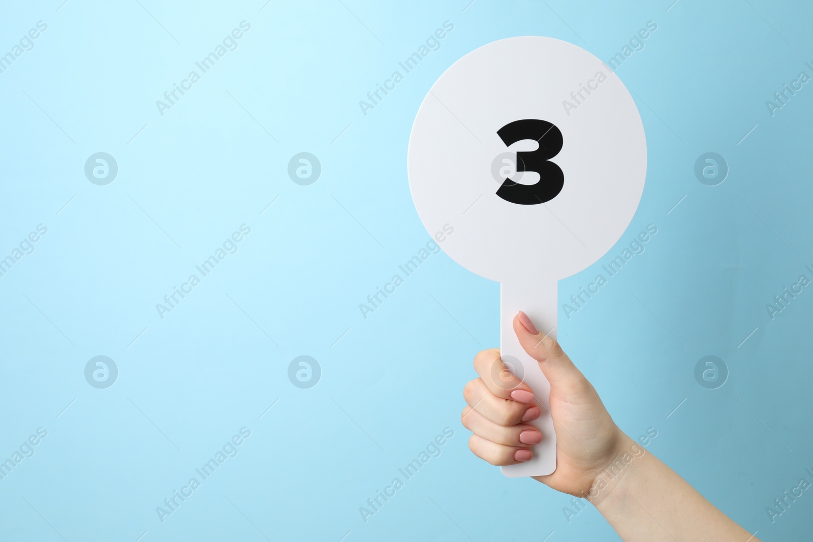Photo of Woman holding auction paddle with number 3 on light blue background, closeup. Space for text