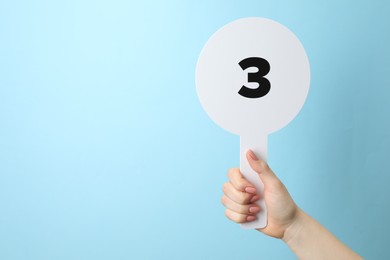 Woman holding auction paddle with number 3 on light blue background, closeup. Space for text