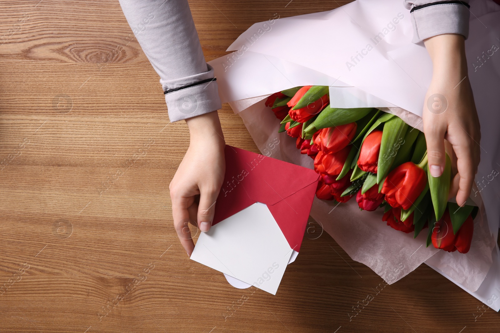 Photo of Woman holding envelope with blank greeting card and bouquet of tulips on wooden table, top view