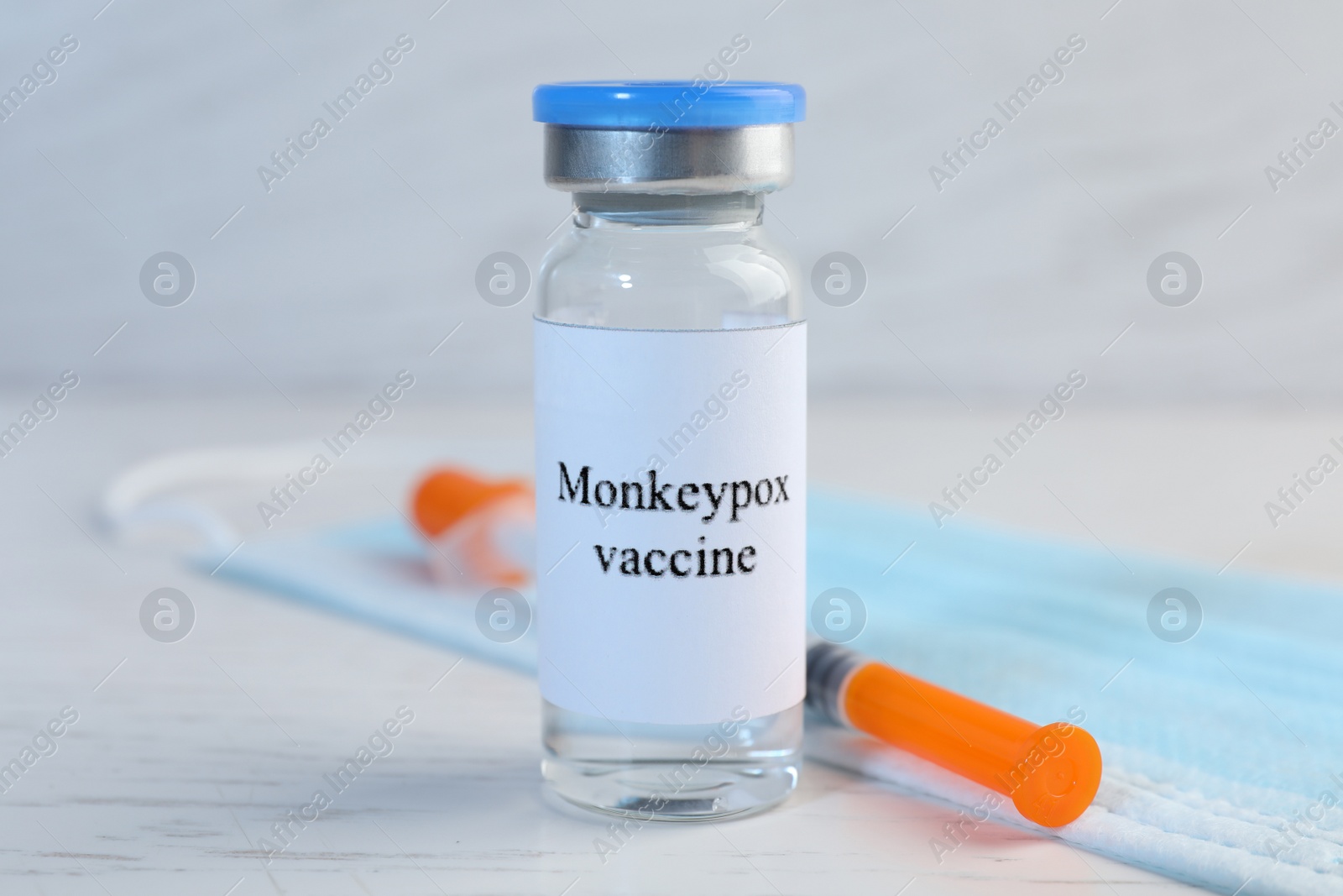 Photo of Monkeypox vaccine in glass vial, medical mask and syringe on white wooden table