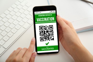 Woman holding smartphone with international certificate of vaccination at white table, closeup