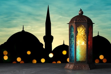 Decorative Arabic lantern on stone surface and silhouette of mosque at sunset on background, space for text