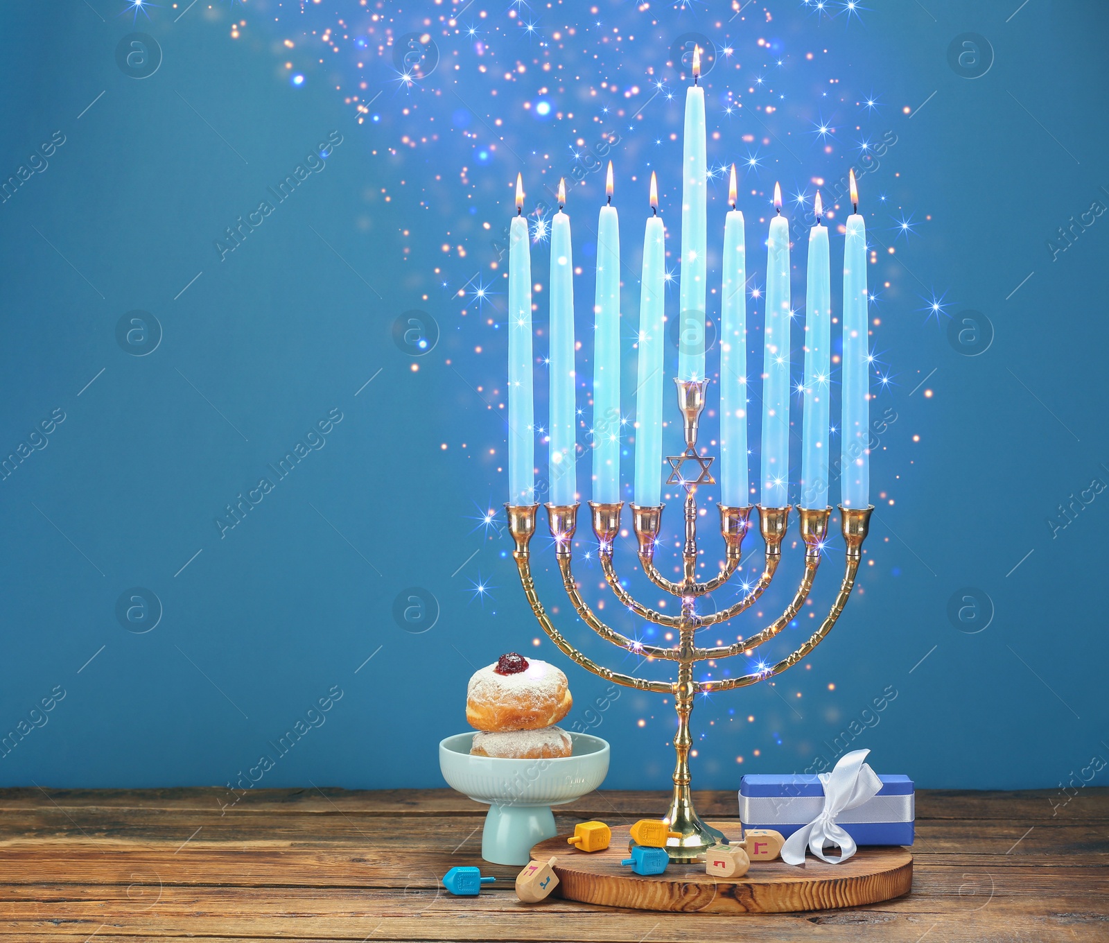 Image of Hanukkah celebration. Menorah with burning candles, dreidels, gift box and donuts on wooden table against light blue background, space for text