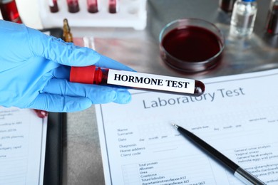 Doctor holding glass tube with blood sample and label Hormone Test at table, closeup